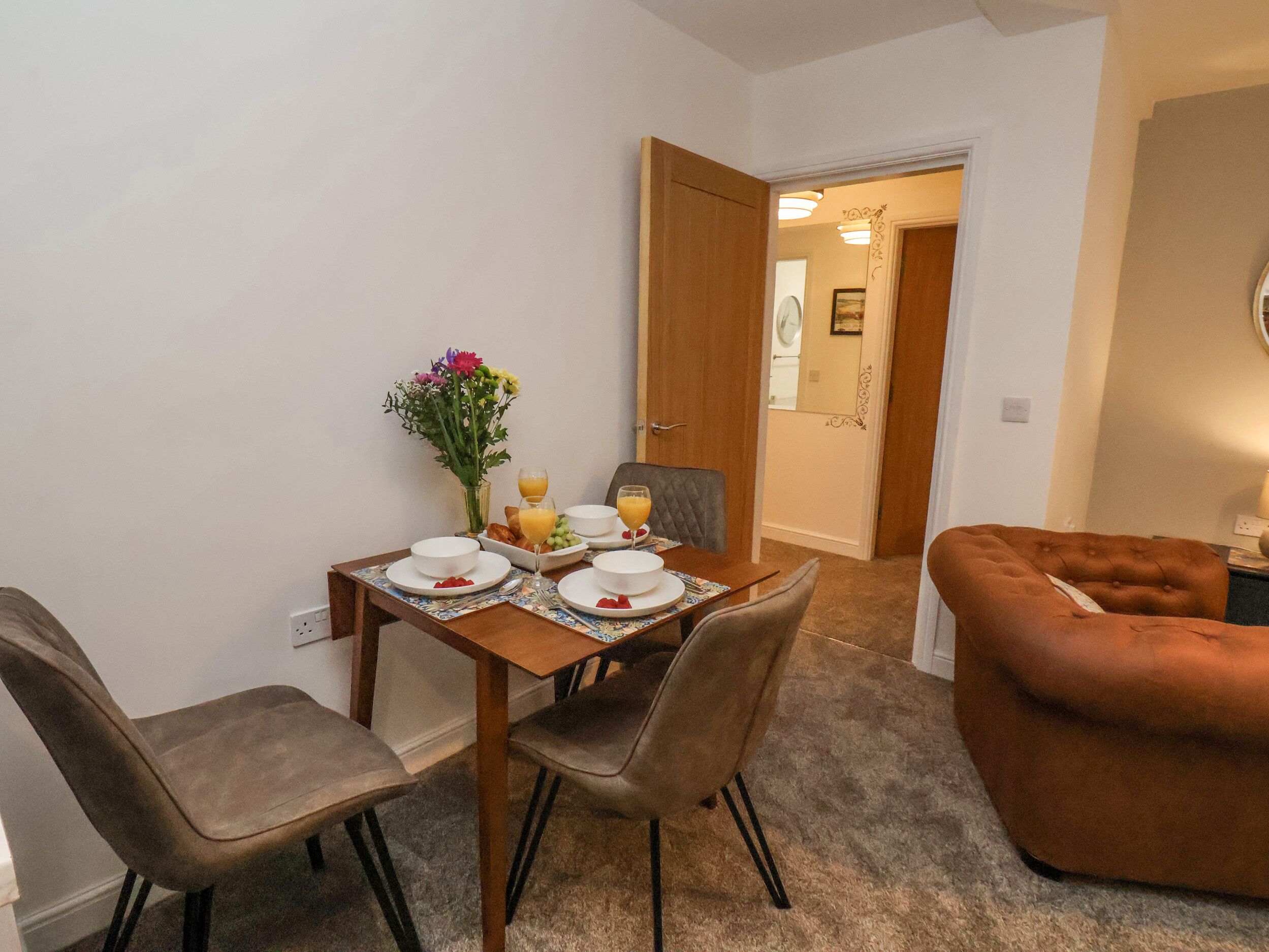 A dining area with a table set for two, two chairs, a vase with flowers, and a breakfast tray with croissants and orange juice.