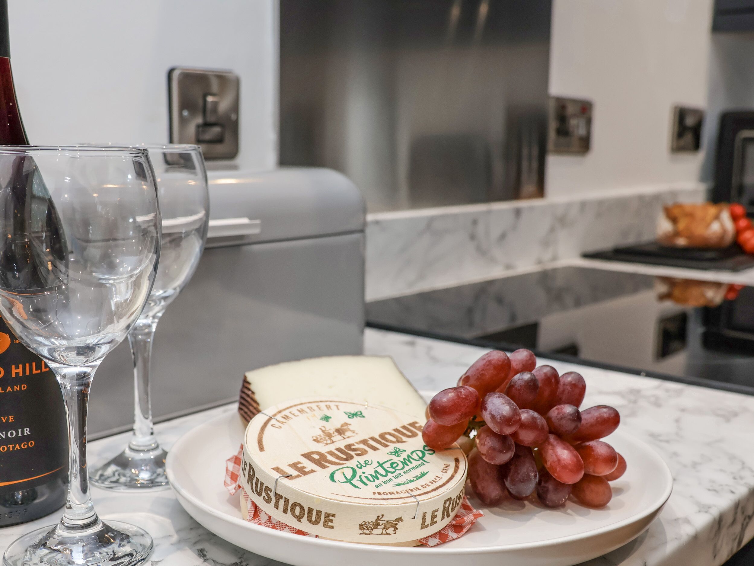 A kitchen counter with a plate of Le Rustique cheese, a bunch of grapes, and two wine glasses.