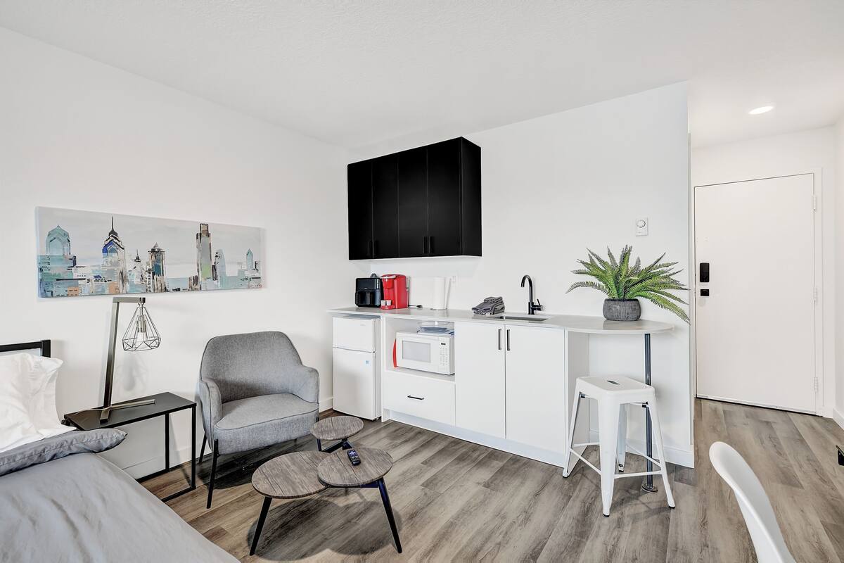 A compact room with a bed, a grey armchair, a small round table, a white kitchenette, a black TV mounted on the wall, and a cityscape painting.