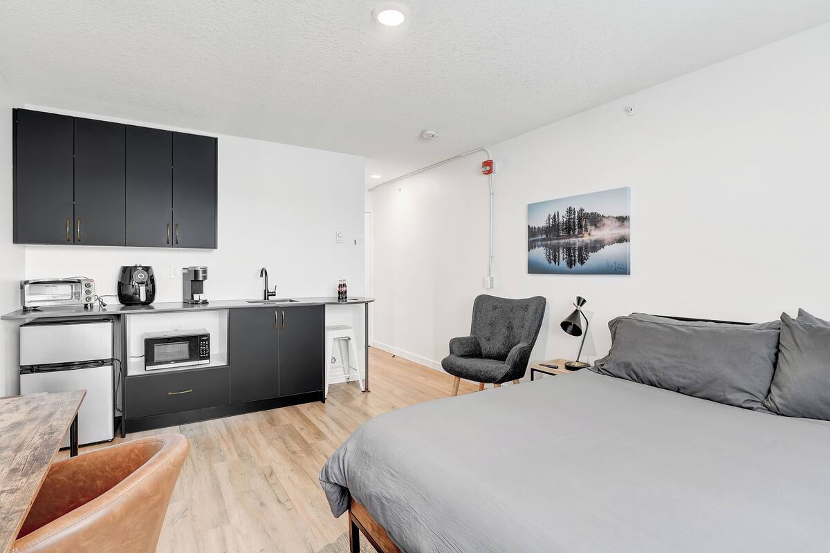 A modern bedroom with a large bed, a desk with a computer, a chair, and a kitchen area with appliances.