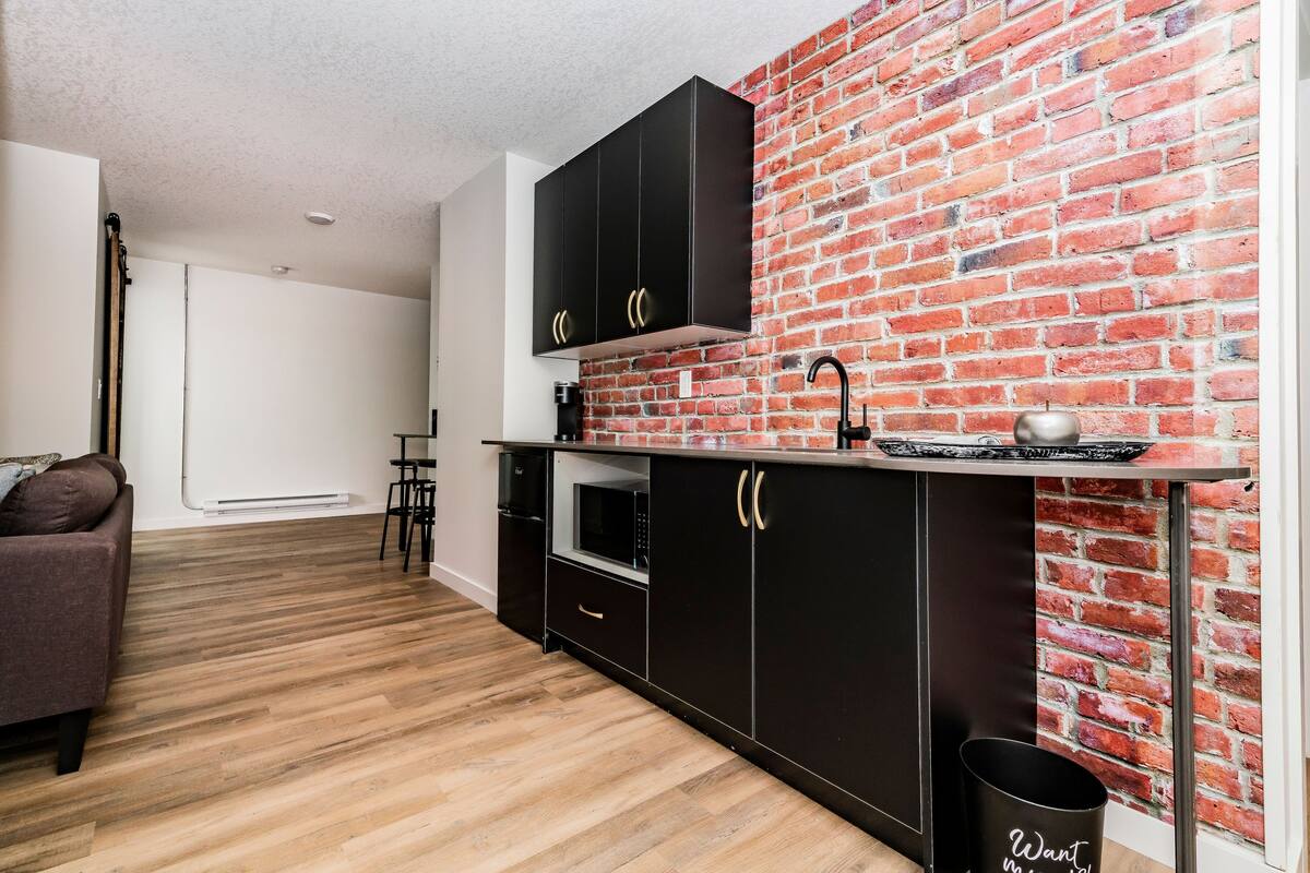 A modern kitchen with black cabinets, a brick wall, and a wooden floor.