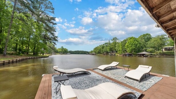 A wooden deck with lounge chairs and a table overlooking a calm body of water, surrounded by trees and a clear sky.