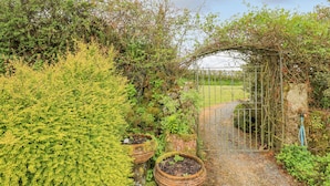A garden with a metal gate, potted plants, and a stone wall. - Gilly Skyber (Helston)