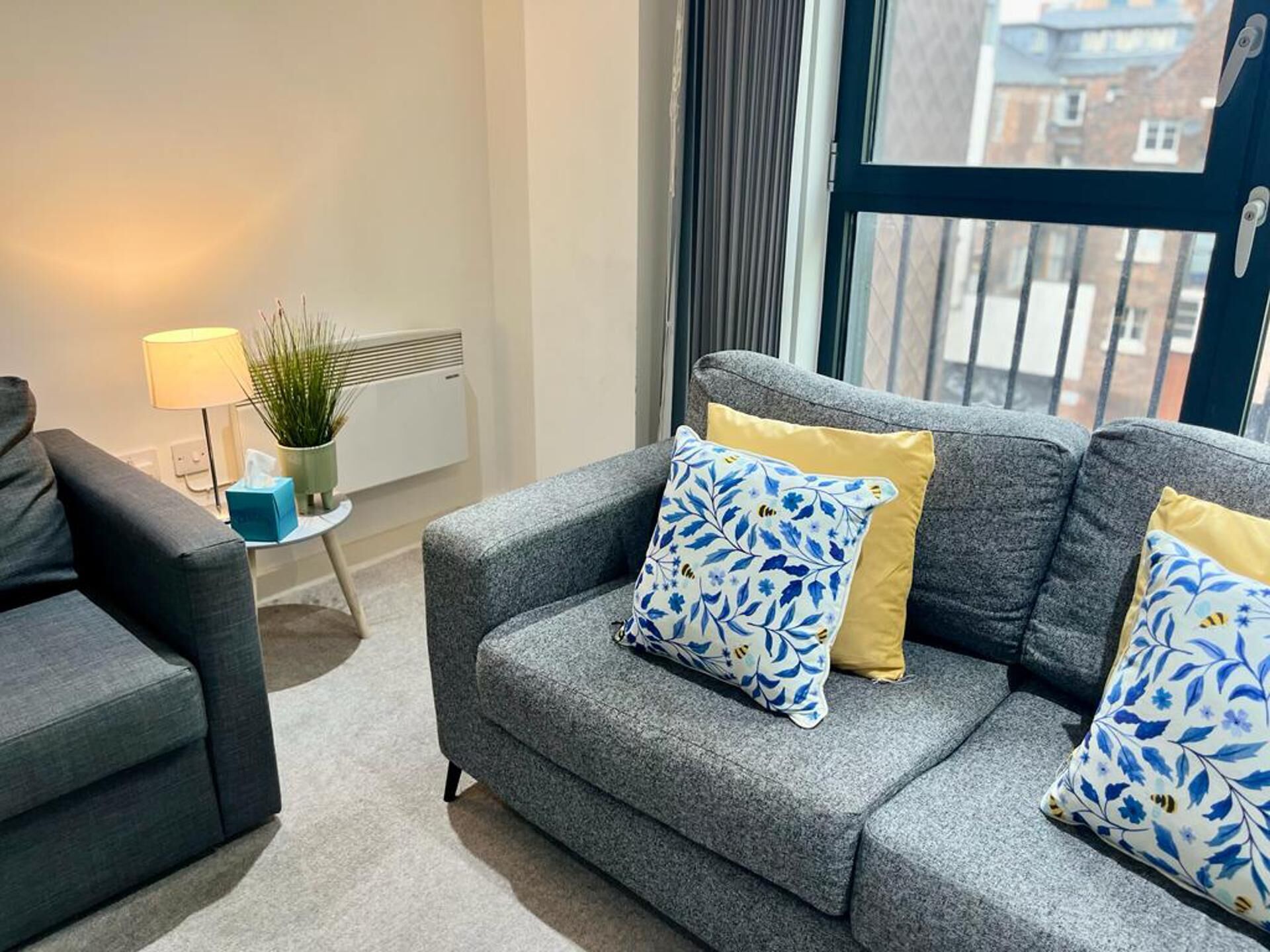 A modern living room with a grey sofa, two patterned cushions, a small side table with a lamp and a plant, and a view of buildings through a window.