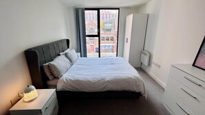 Ein modernes Schlafzimmer mit einem großen Bett, einem Nachttisch mit Lampe, einem Kleiderschrank und Blick auf Gebäude durch ein Fenster.