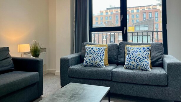 Ein modernes Wohnzimmer mit einem grauen Sofa, zwei gemusterten Kissen, einem kleinen Tisch, einer Lampe und Blick auf Gebäude durch ein großes Fenster.