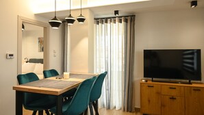Zona pranzo moderna con tavolo in legno, sedie color petrolio e illuminazione a sospensione. Un televisore a schermo piatto è montato su un mobile in legno.
