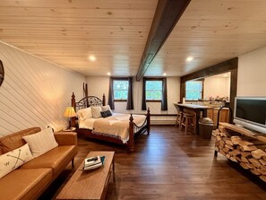Une piĂšce confortable avec un lit, une table basse en bois, un tĂ©lĂ©viseur posĂ© sur un meuble en bois, et un coin cuisine Ă©quipĂ© dâun Ă©vier et de quelques tabourets.