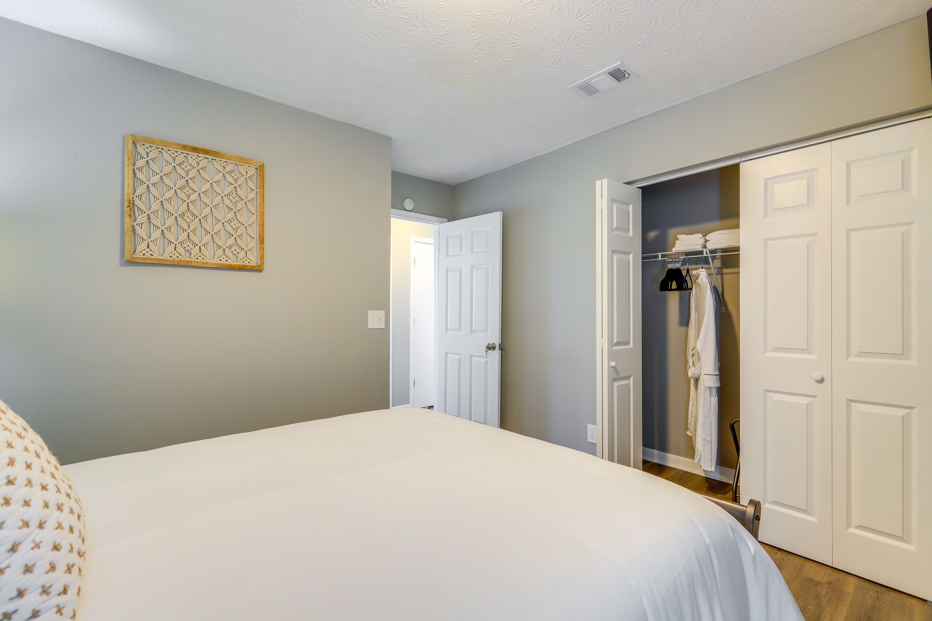 A bedroom with a large bed, a closet with hanging clothes, and a door leading to another room.