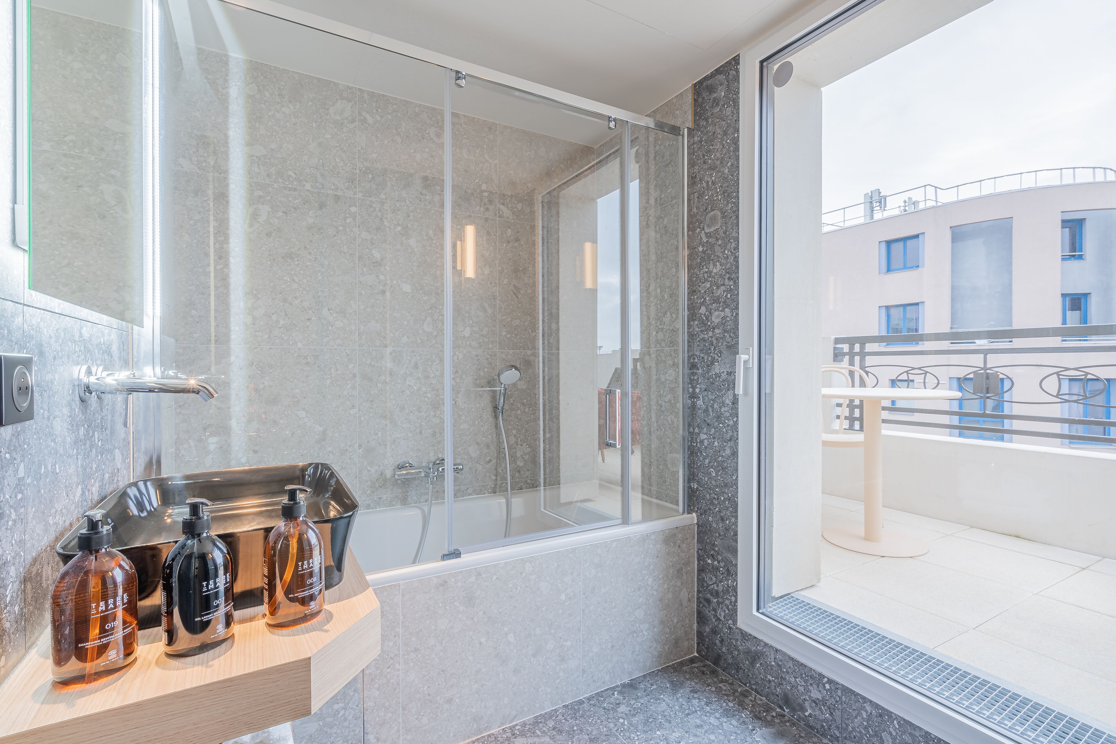 Een moderne badkamer met een douche in een glazen cabine, een wastafel met flessen en uitzicht op het omliggende gebouw.