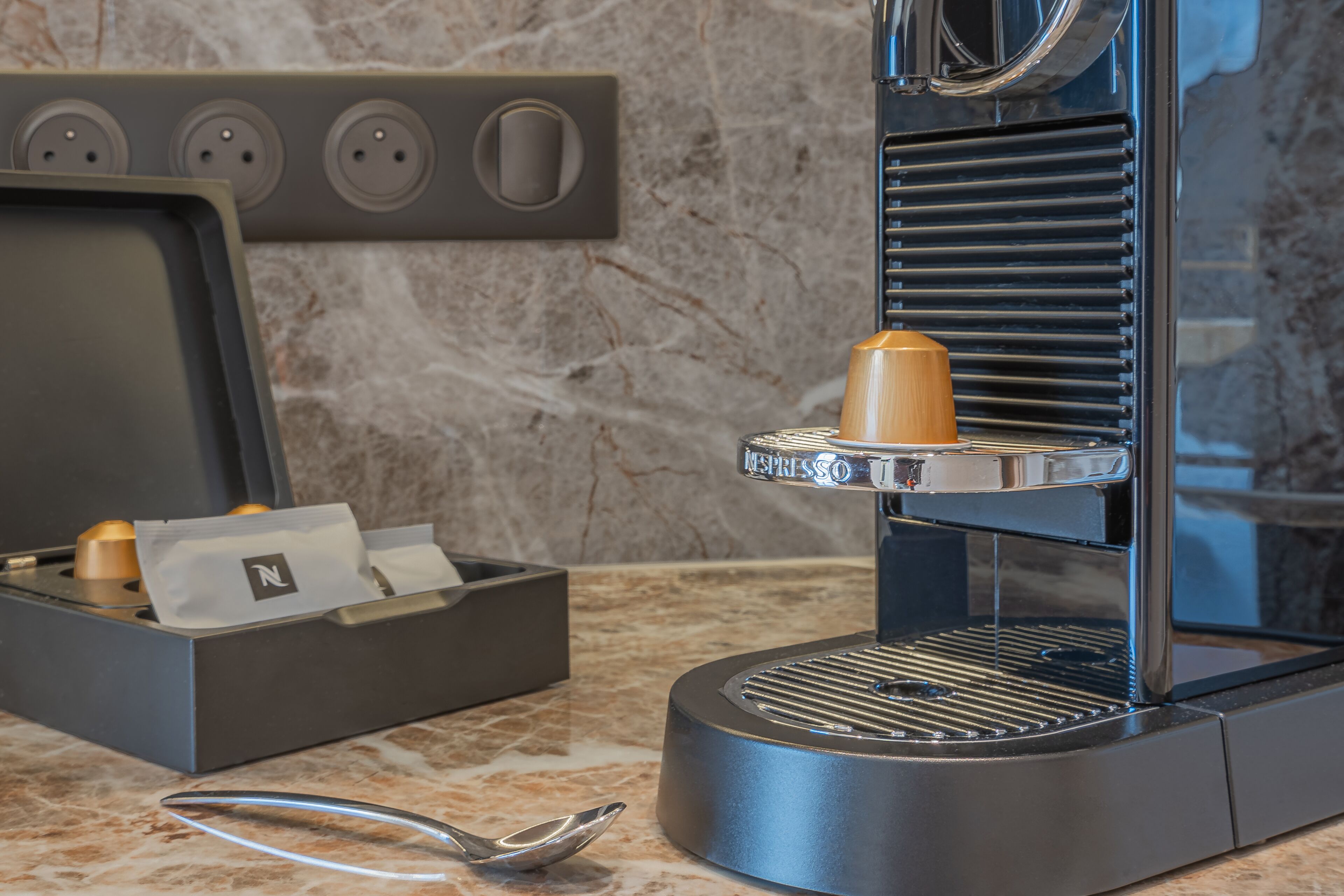 Una moderna macchina da caffè espresso con macinacaffè integrato, portabicchieri per capsule e un cucchiaino di metallo su un piano di marmo.