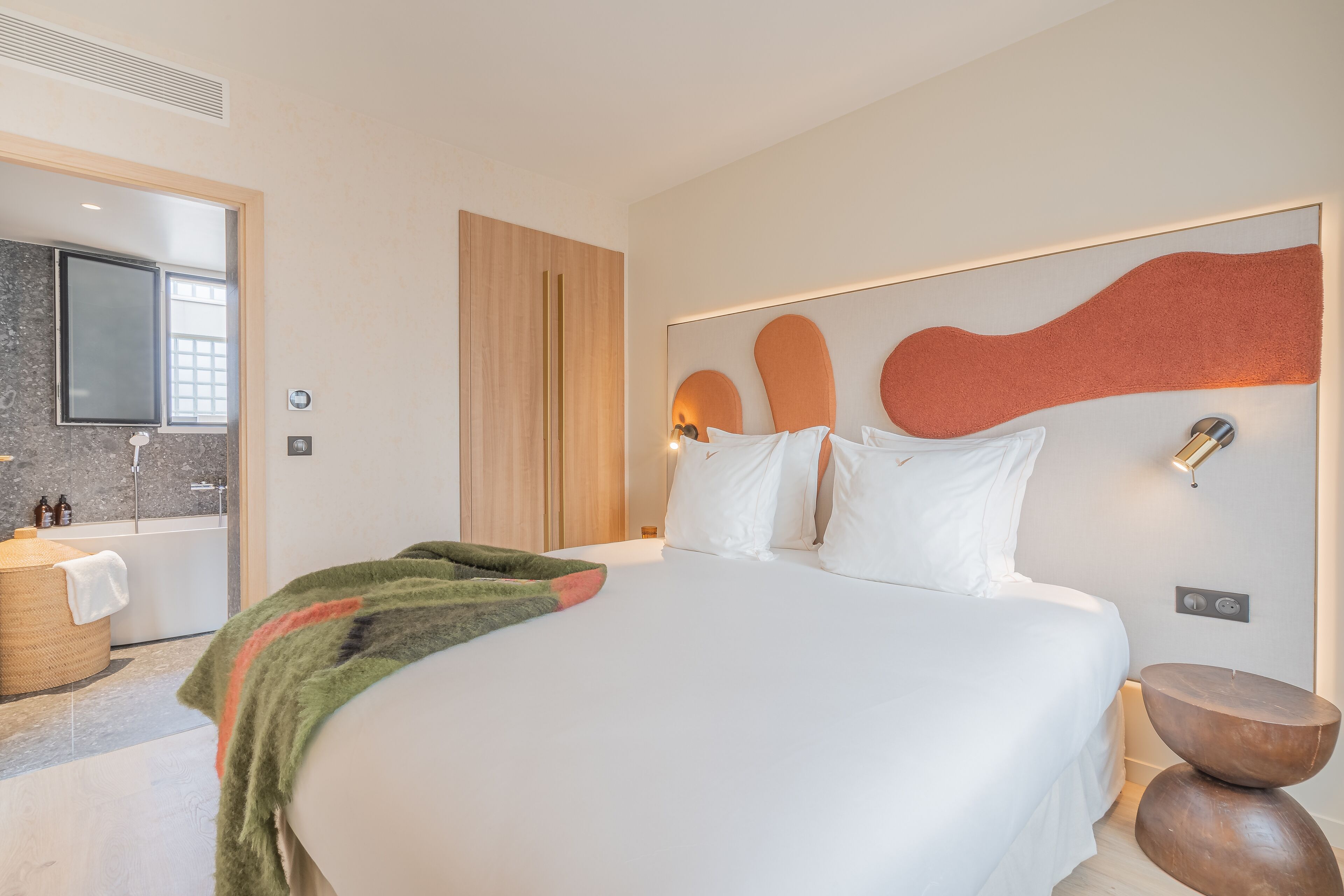 A modern hotel room with a large bed, a wooden headboard, a wall-mounted TV, a bathroom with a bathtub, and a round wooden side table.