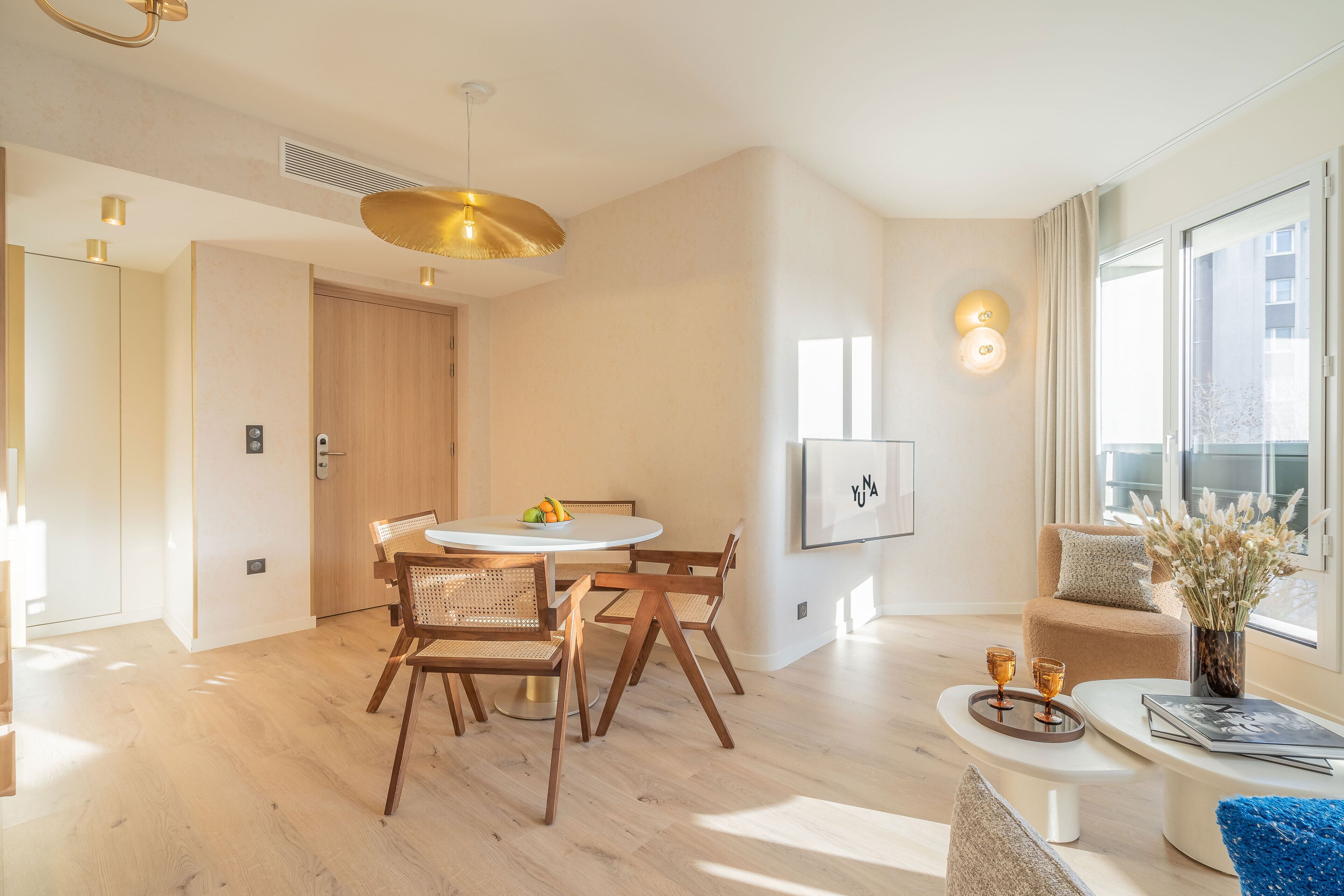 Uma sala de estar moderna com uma mesa de jantar redonda, cadeiras de madeira, um sofá e uma janela grande com vista para edifícios.