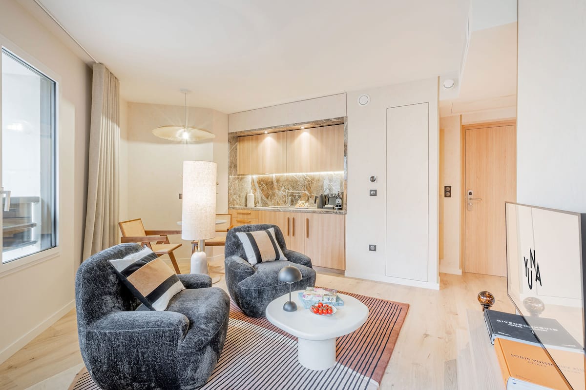 Ein modernes Wohnzimmer mit zwei grauen Sesseln, einem weißen Couchtisch und einem gestreiften Teppich. Im Hintergrund ist eine Küche mit Holzschränken und einer Marmor-Spritzschutzwand zu sehen.