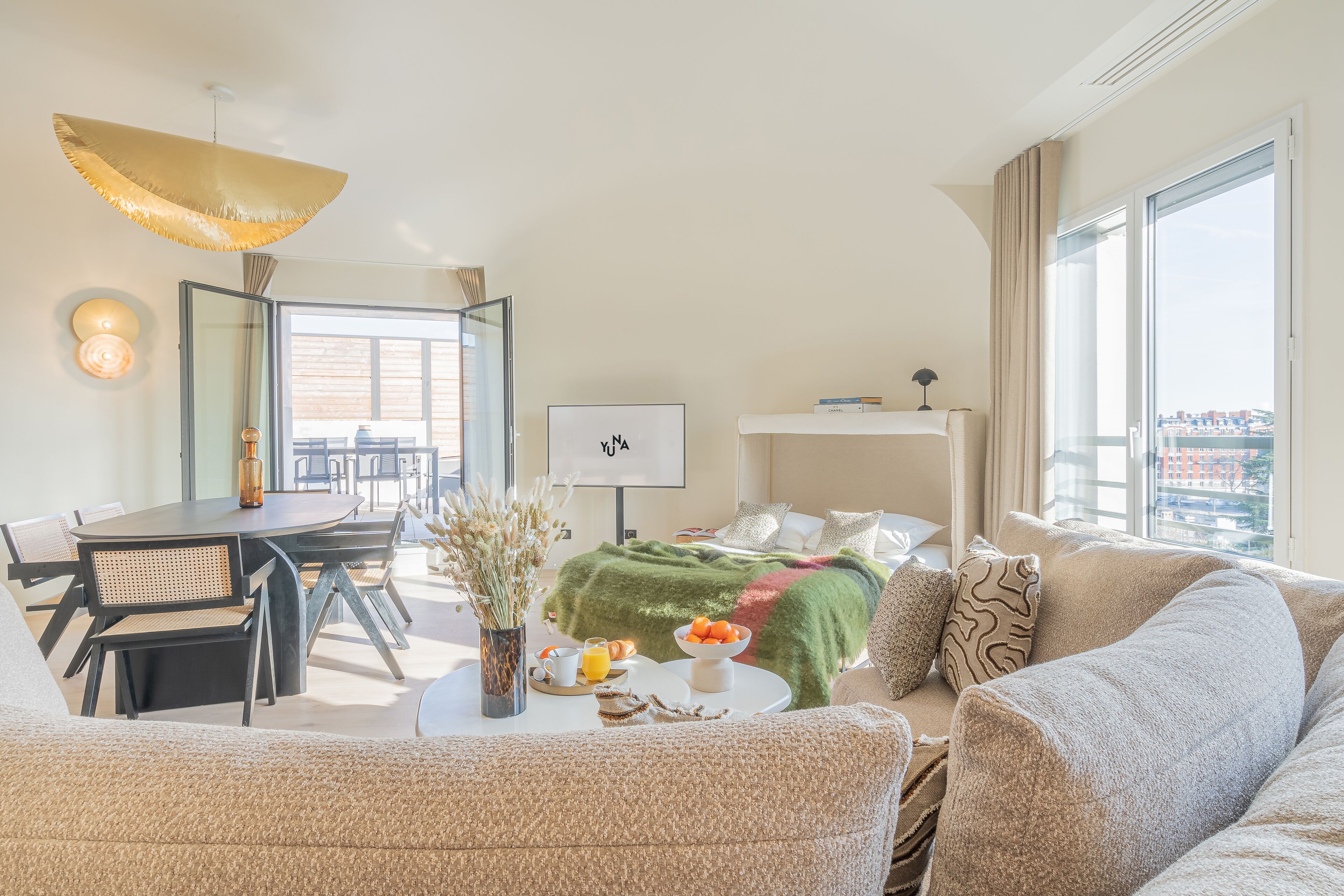 A modern hotel room with a dining area, a bed with a green blanket, a sofa, and a large window with a city view.