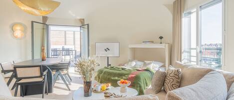 A modern hotel room with a dining area, a bed with a green blanket, a sofa, and a large window with a city view.