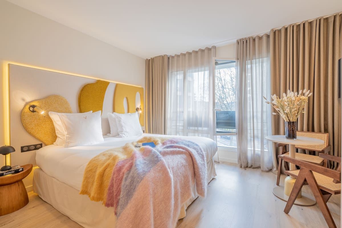 Ein modernes Hotelzimmer mit einem großen Bett, einem Nachttisch mit Lampe, einem kleinen runden Tisch mit Stuhl und Blick ins Freie durch das Fenster.