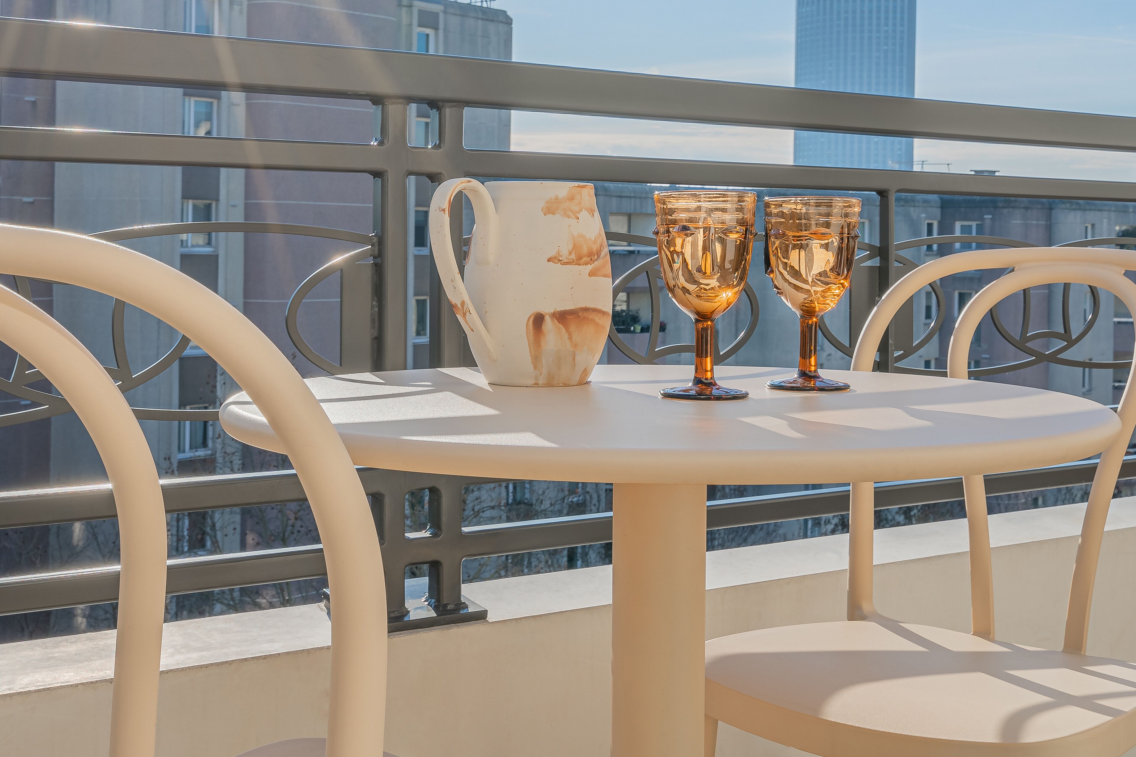 A round table with two glasses and a pitcher on a balcony with metal railings and city views.