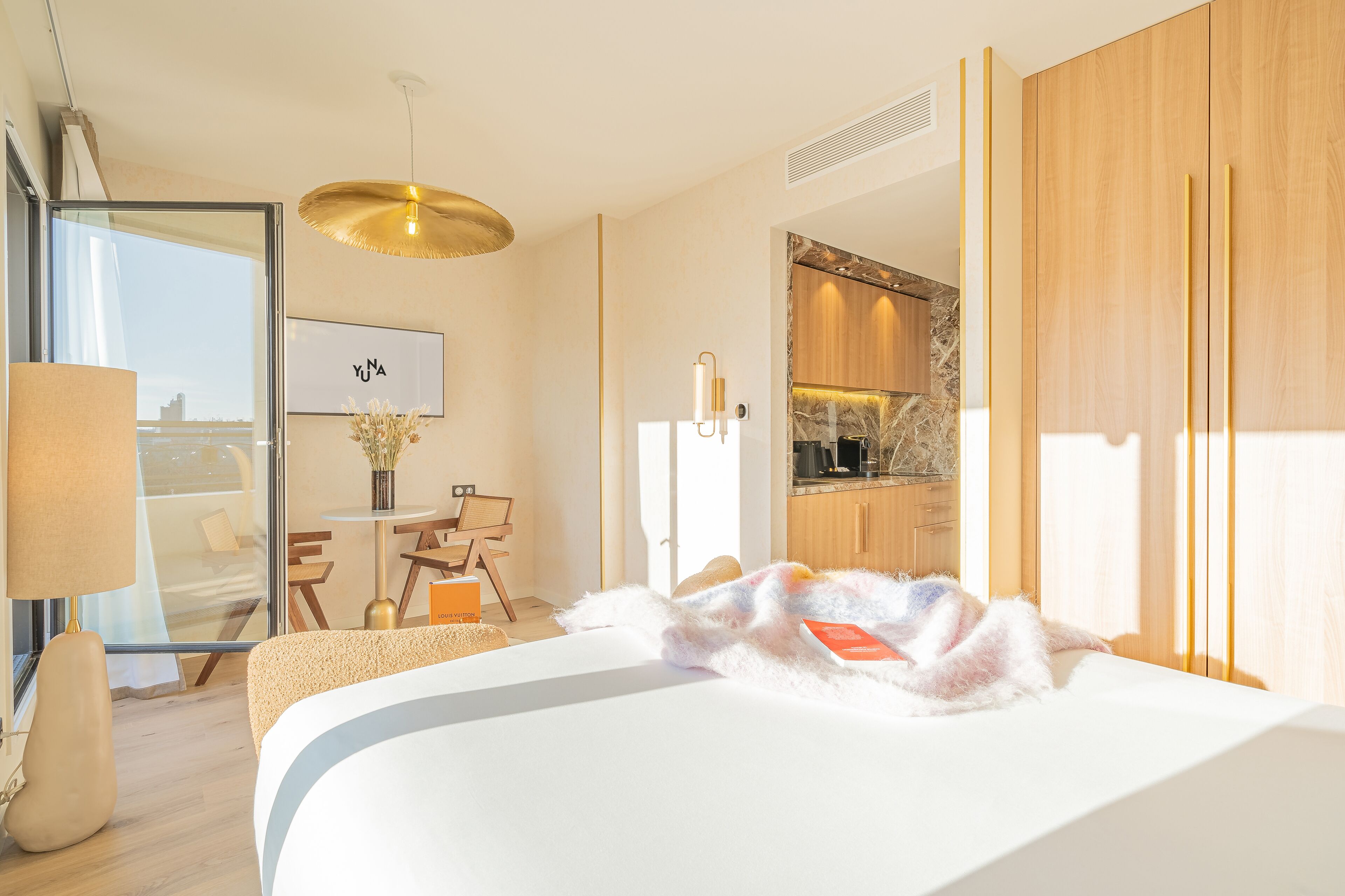 Quarto de hotel moderno com uma cama grande, área de refeições com mesa e cadeiras, cozinha e vista através de uma porta de vidro.