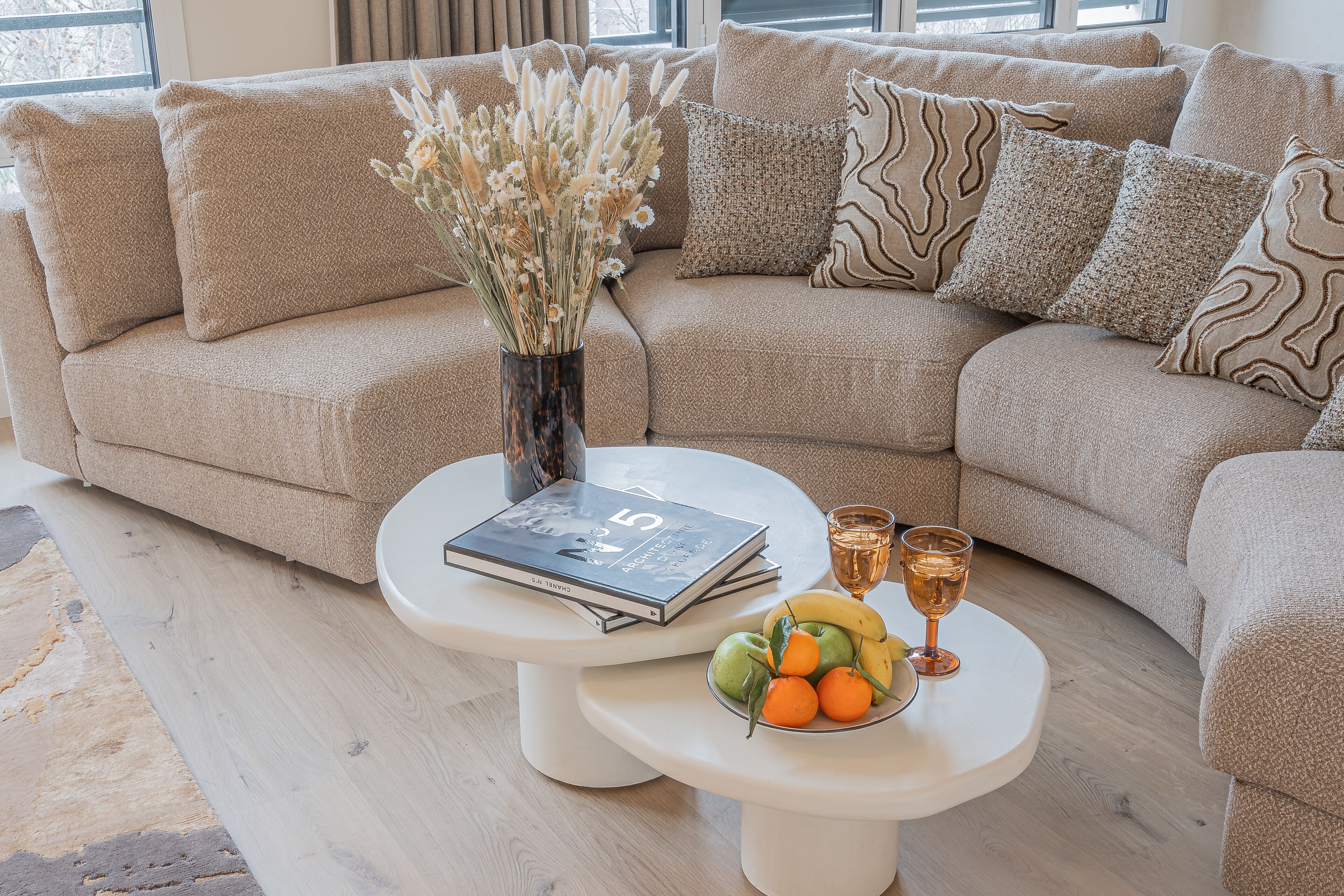 A beige sofa with patterned pillows, a vase with dried flowers, a magazine, and a fruit bowl with glasses on a white coffee table.