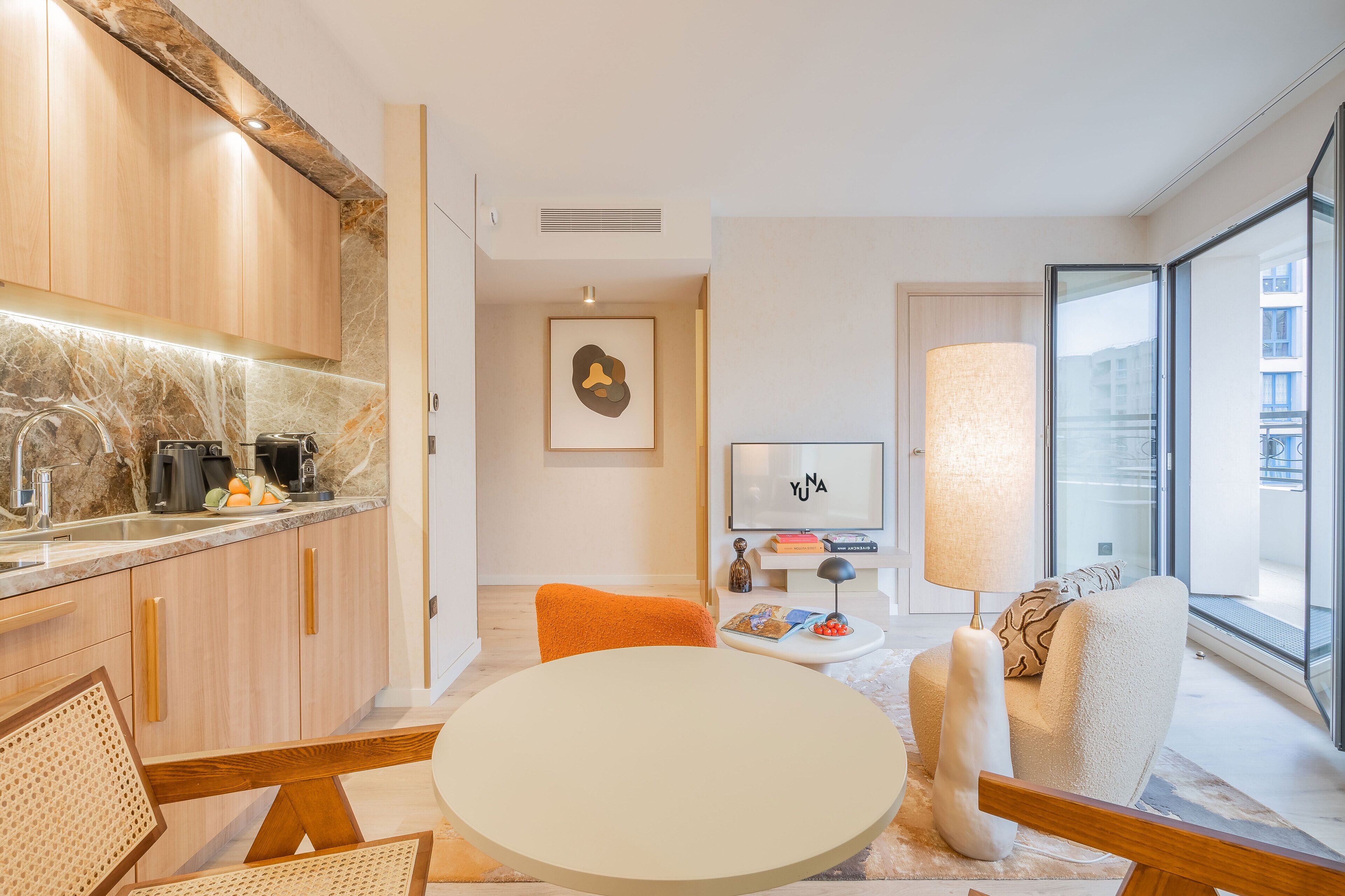 A modern kitchen with marble countertops, wooden cabinets, and a dining area with a round table and orange chairs.