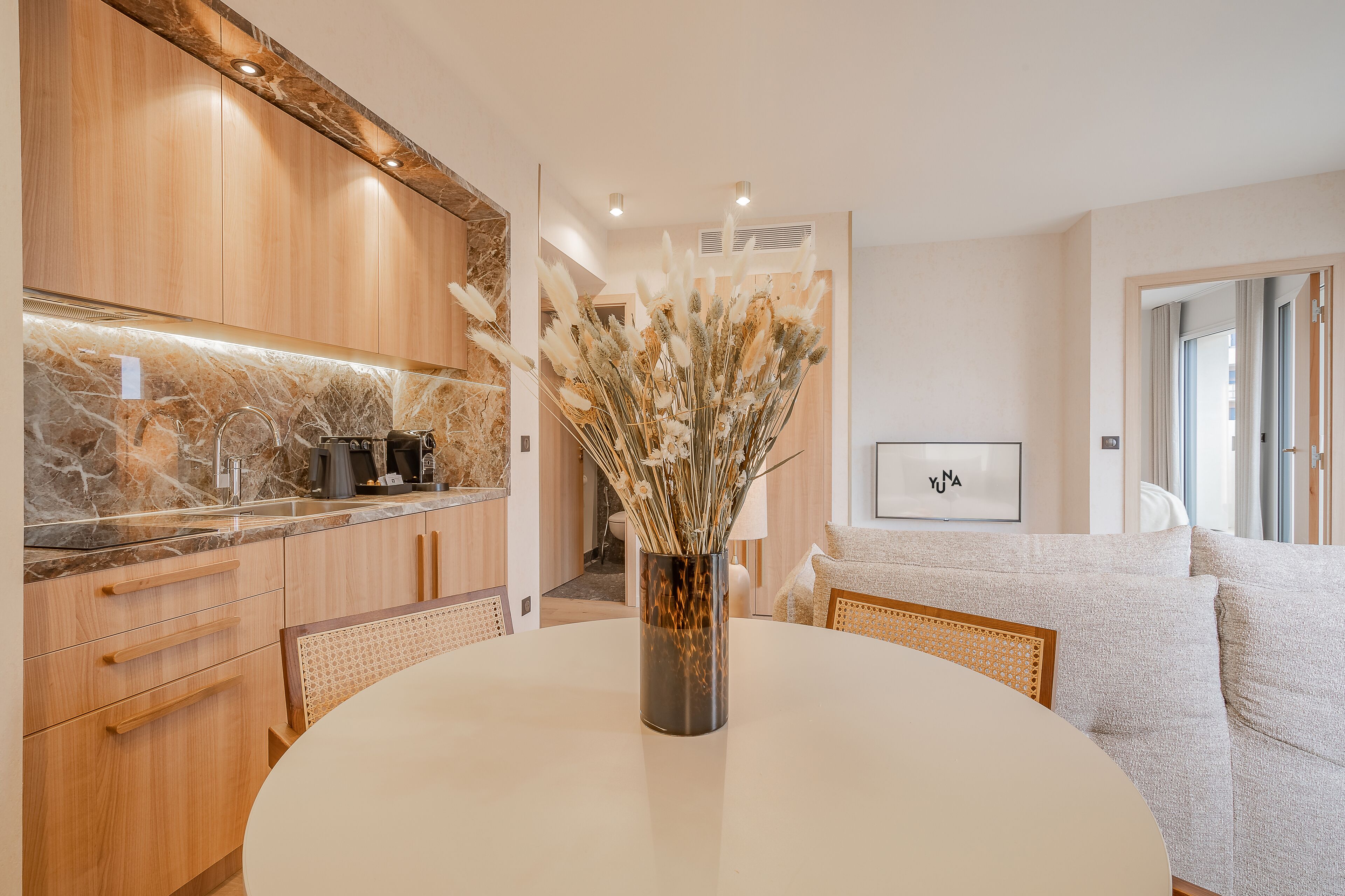 Een moderne keuken met houten kastjes, een marmeren aanrechtblad en een vaas met gedroogde bloemen op een ronde tafel.