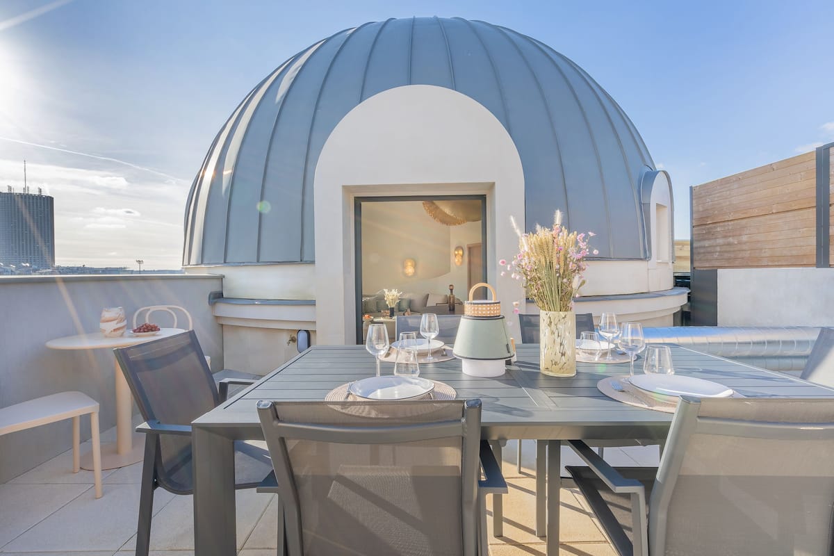 Ein Essbereich im Freien mit Tisch für zwei, einer großen Kuppelstruktur und Blick auf die Stadtlandschaft.