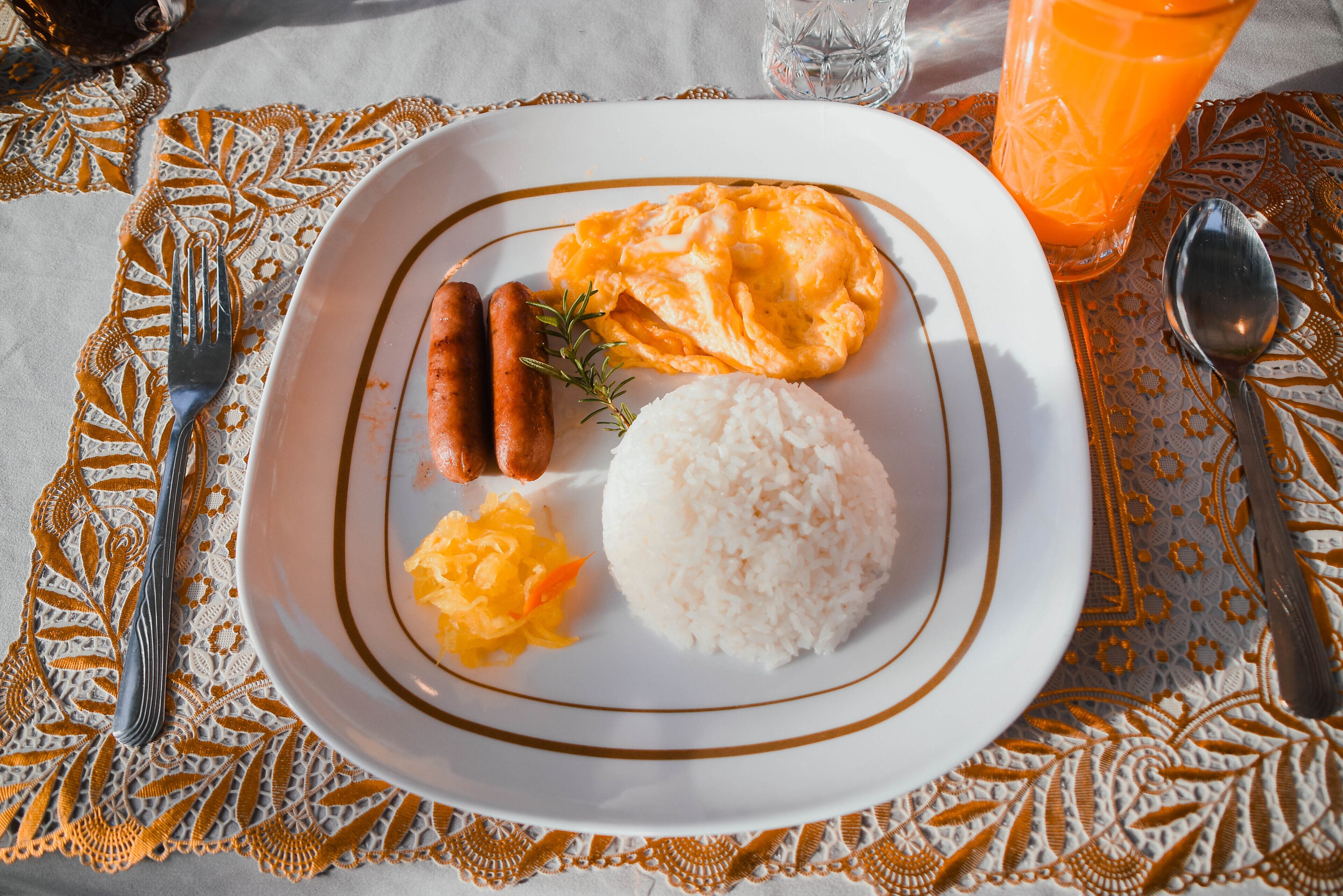 A plate with scrambled eggs, sausages, rice, and sauerkraut on a table with a lace tablecloth.