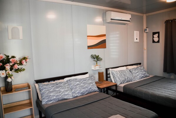 Habitación de hotel con dos camas, una mesita de noche de madera, un jarrón de flores y un aire acondicionado instalado en la pared.