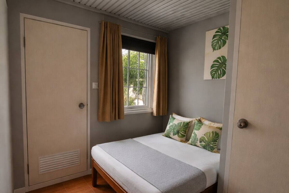 A bedroom with a bed, a window with curtains, a door, and a wall with a plant.