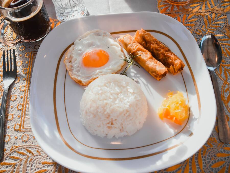 A plate with fried egg, rice, spring rolls, and orange slices on a table with a lace tablecloth.