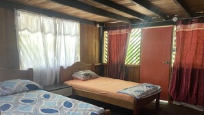 A room with two beds, wooden headboards, a window with sheer curtains, and a door with patterned curtains. - CABAÑAS TAKANA (Leticia)