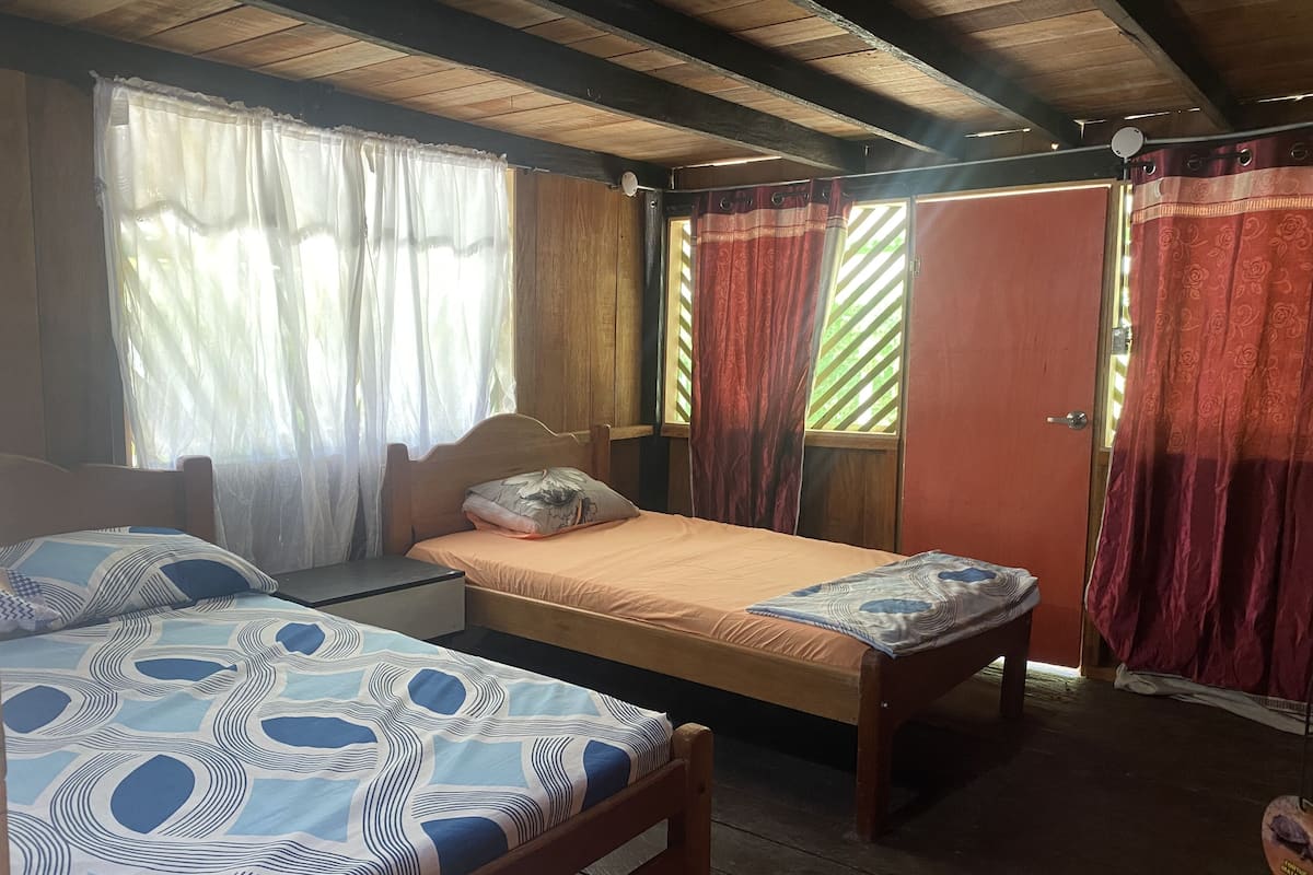 A room with two beds, wooden headboards, a window with sheer curtains, and a door with patterned curtains.