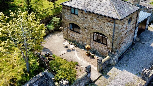 A stone building with a tiled roof, arched windows, and a small garden area with a table and chairs. - The Coach House (Holywell)