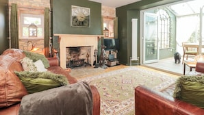 A cozy living room with a fireplace, leather sofas, a patterned rug, and a large window offering a view of the outdoors. - Yarn Cottage (Broadway)