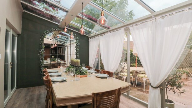 A dining area with a glass enclosure, wooden furniture, and a table set for four. - Yarn Cottage (Broadway)