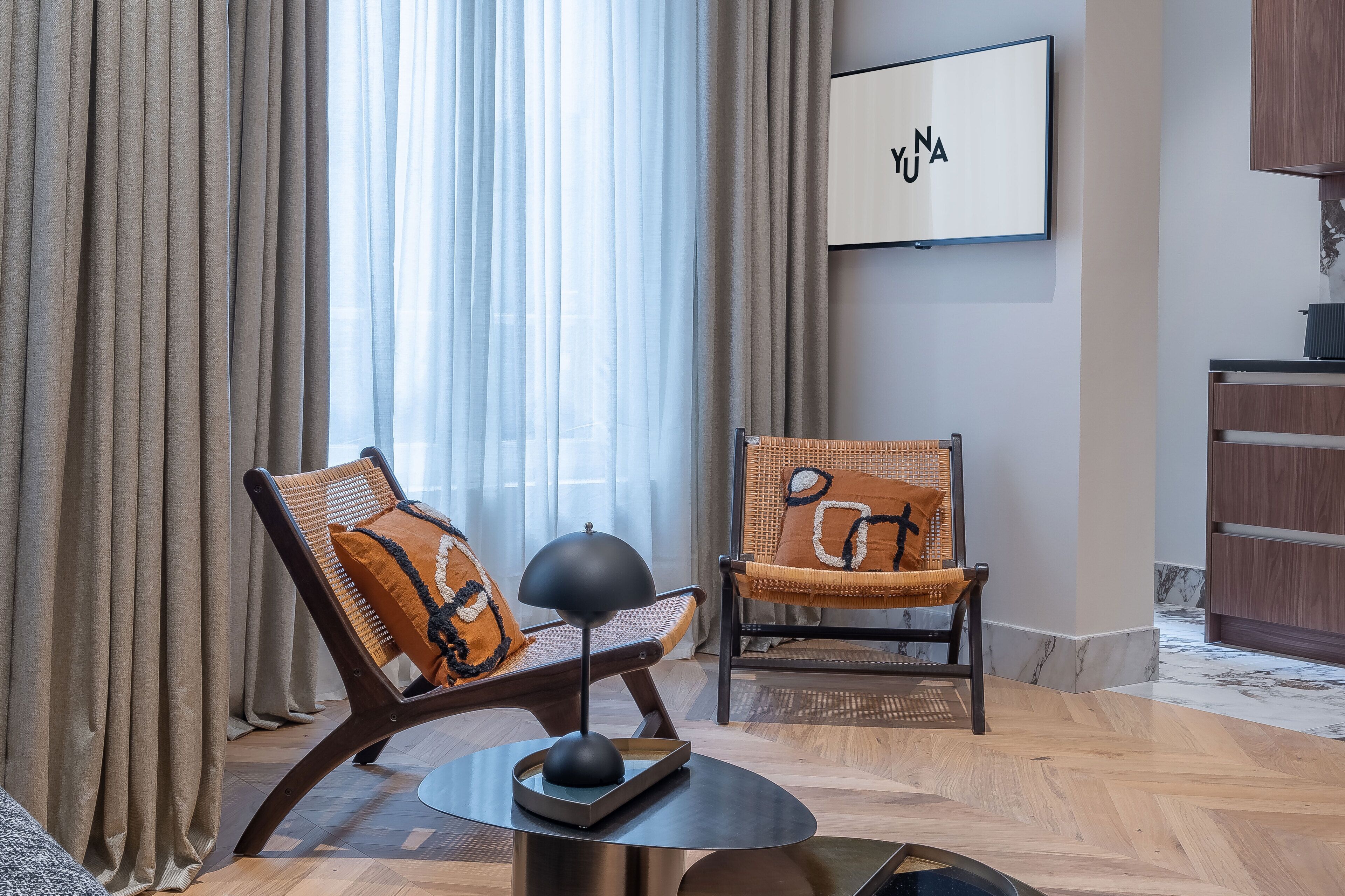 Een moderne woonkamer met twee rieten fauteuils, een ronde salontafel en een aan de muur bevestigde televisie.