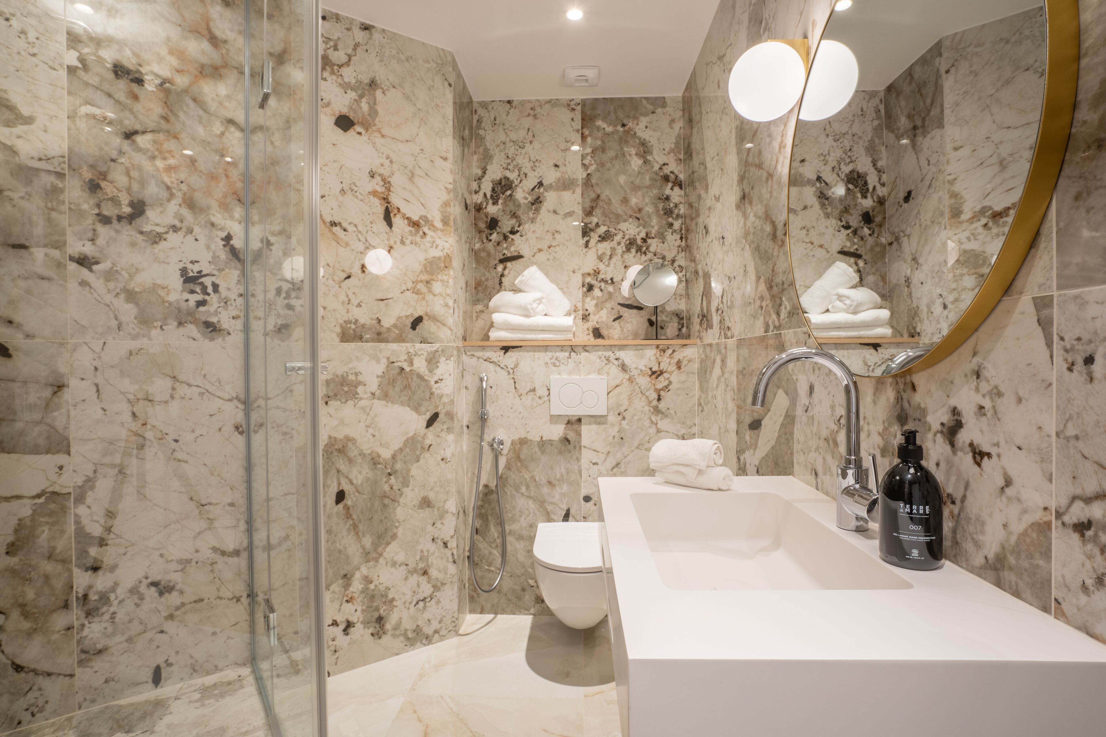 Een moderne badkamer met een marmeren wand, een glazen douchecabine, een witte wastafel en een spiegel met een gouden lijst.