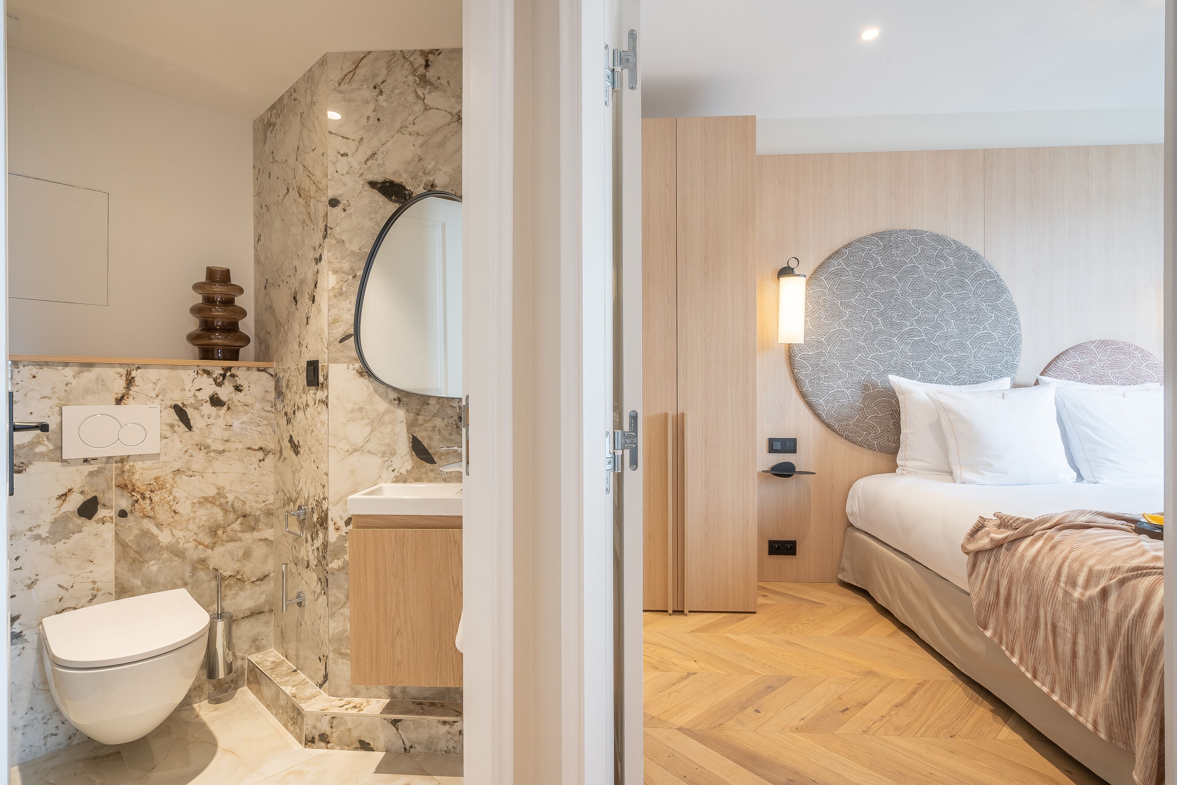 A modern bathroom with a marble wall, a toilet, and a sink. The room has a wooden floor and a bed with a circular headboard.
