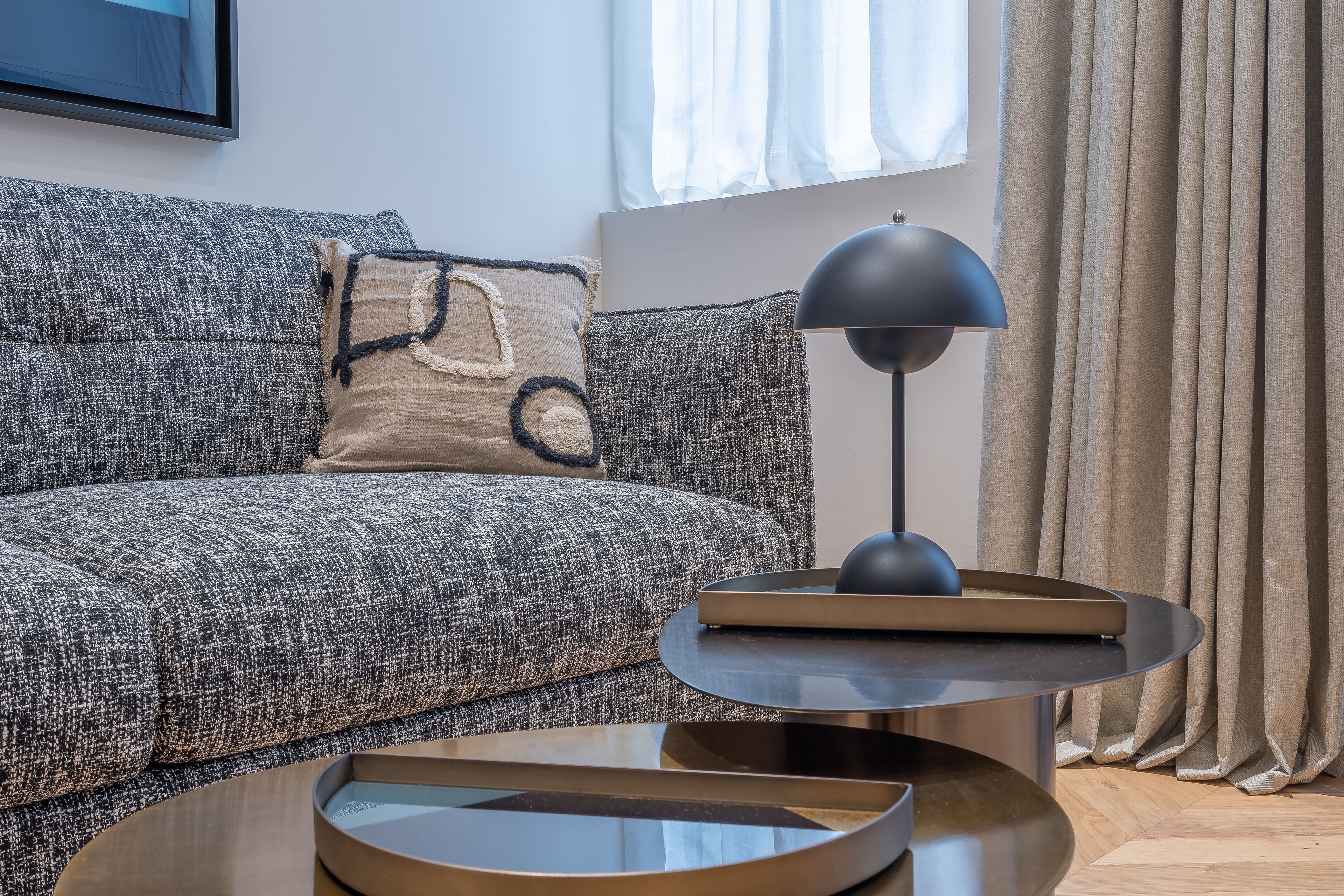 Una sala moderna con un sofá gris con textura, una mesa redonda, una lámpara de pie negra y un cojín beige con diseño geométrico.