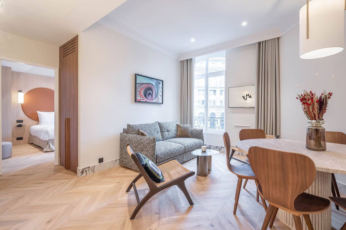 Ein modernes Wohnzimmer mit einer Couch, einem Esstisch mit Stühlen und einem großen Fenster mit Blick auf Gebäude.