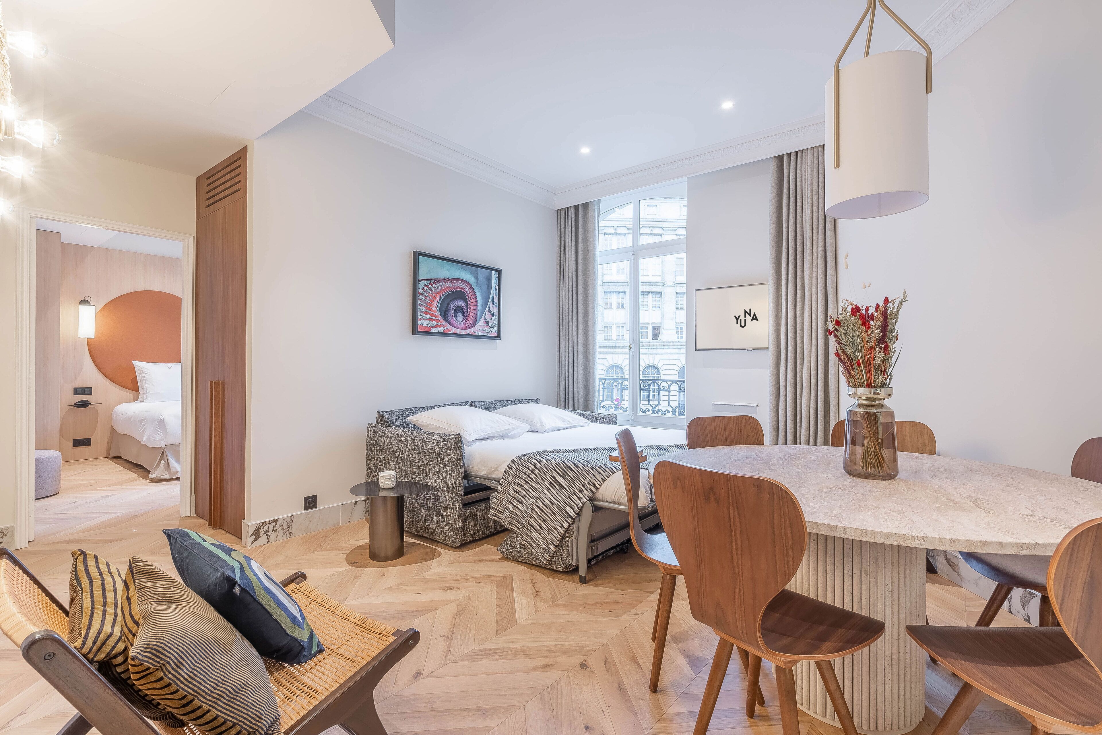 Moderni hotellihuone, jossa on sänky, ruokapöytä ja tuolit. Huoneessa on ikkuna, jossa on verhot, ja seinällä on taulu.