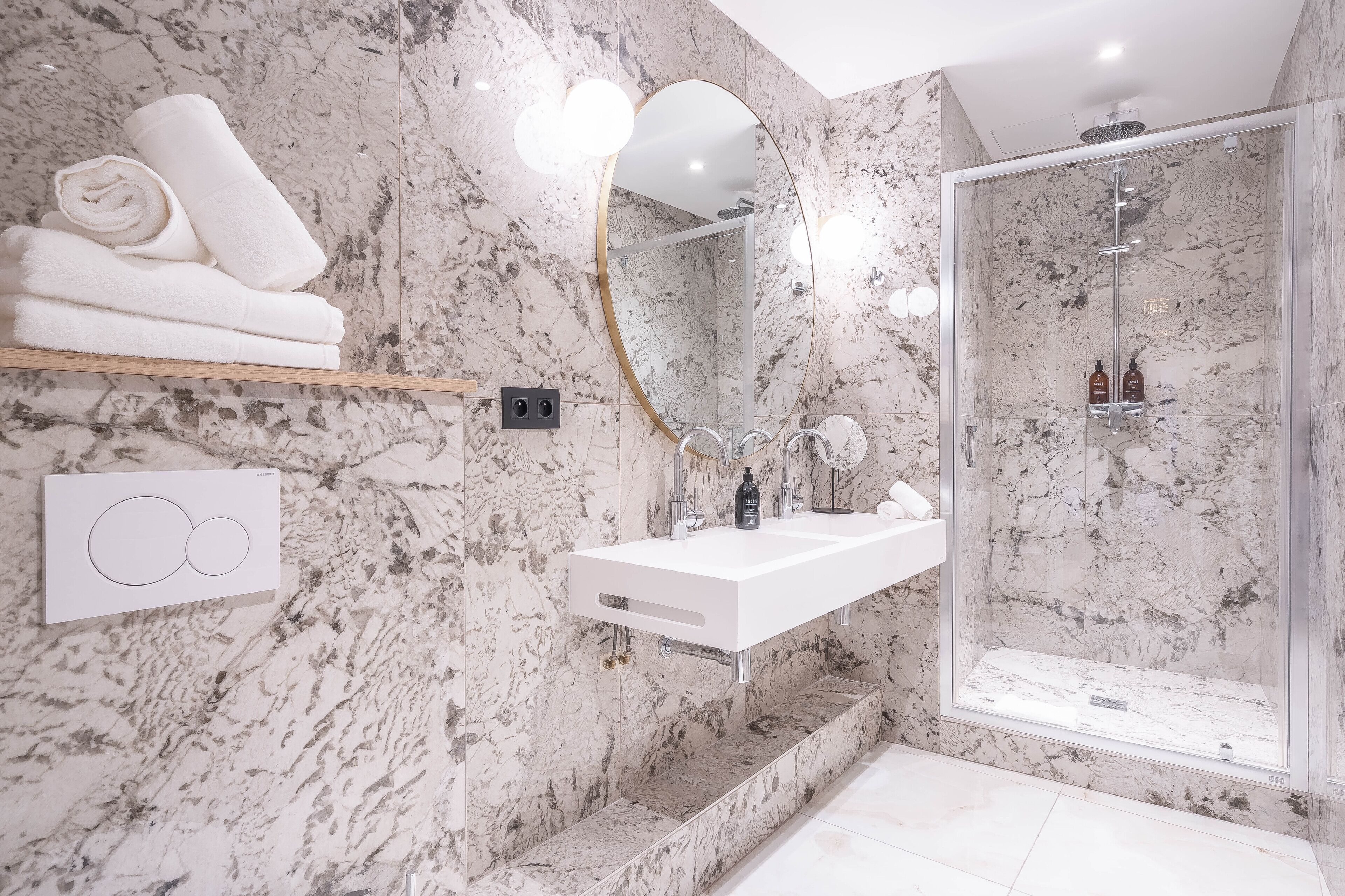 Une salle de bain moderne avec un mur en marbre, un lavabo blanc, une douche en verre et des serviettes soigneusement pliées sur une étagère.