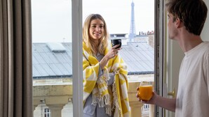 Una persona sosteniendo un teléfono inteligente y un vaso de jugo de naranja, de pie junto a una ventana con vistas a edificios y una torre.
