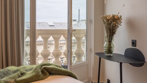 Habitación de hotel con un ventanal grande, una cama con una manta verde, un florero con flores y una mesita.