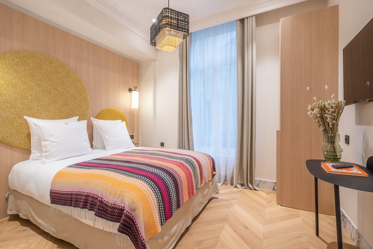 Ein Hotelzimmer mit einem großen Bett, einem Nachttisch mit Blumen in einer Vase und einem großen Fenster mit Vorhängen.