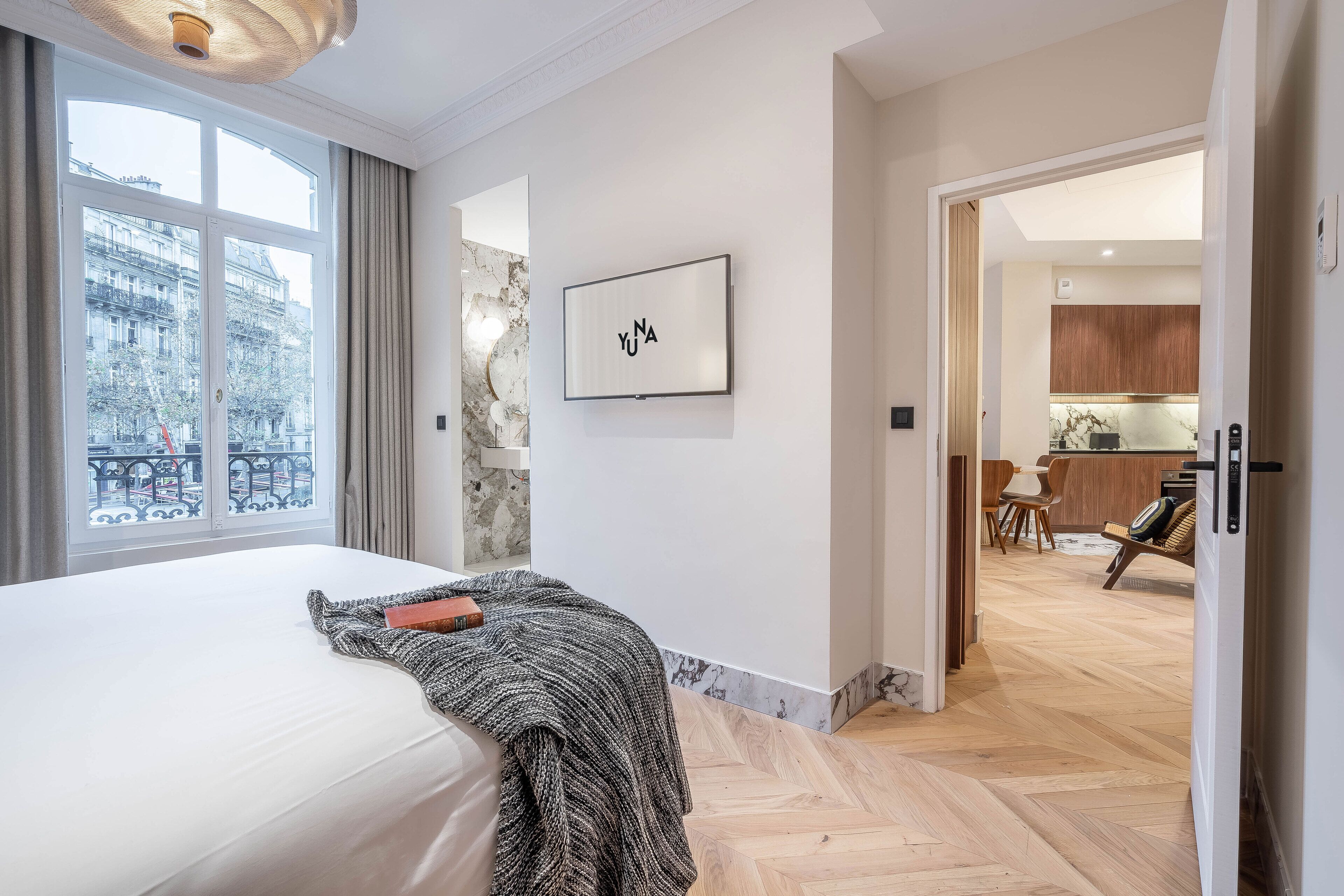 Une chambre d’hôtel avec un grand lit, une vue sur la ville par la fenêtre, une œuvre d’art encadrée au mur et un plancher en bois.