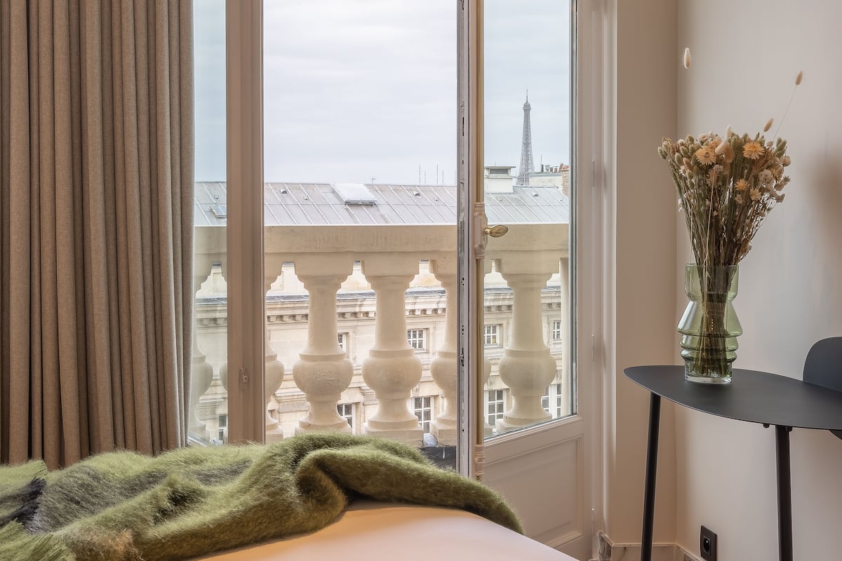 Ein Hotelzimmer mit großem Fenster, einer grünen Tagesdecke, einer Vase mit getrockneten Blumen und Blick auf ein Gebäude mit einem Turm.