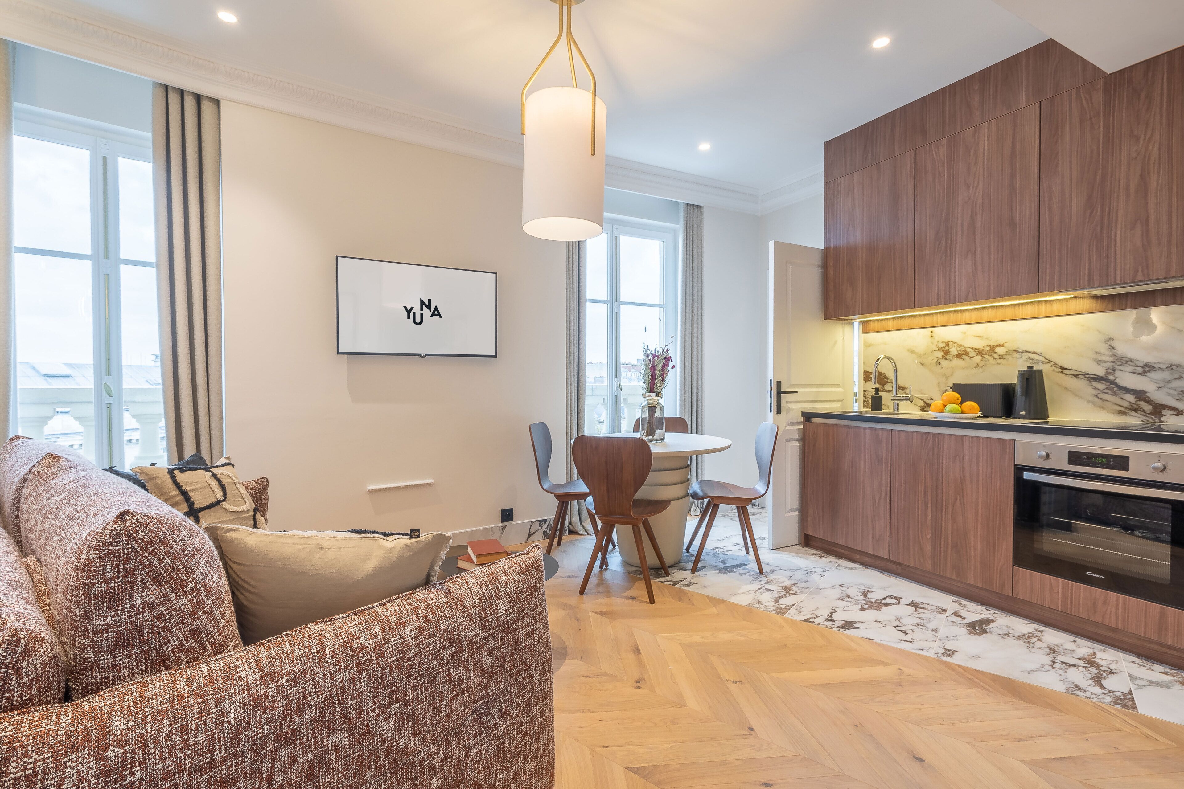 Una sala de estar moderna con un sofá, una mesa de comedor y sillas. Cuenta con una zona de cocina con armarios de madera y encimera de mármol.