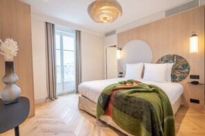 Une chambre d’hôtel moderne avec un grand lit, une tête de lit ronde, une table de chevet agrémentée d’un vase, et une vue sur la ville par la fenêtre.